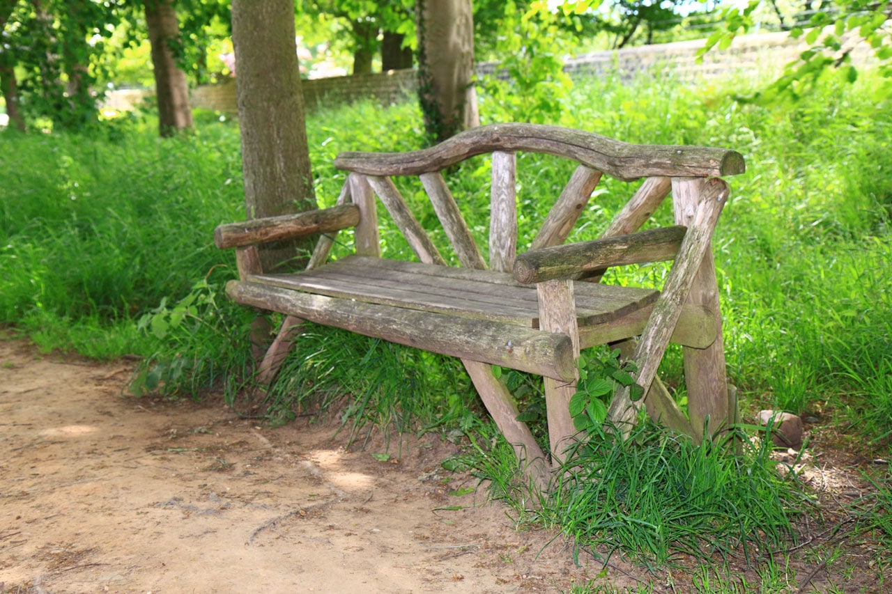 Banc de jardin tendance en vieux bois avec une grande planche et plusieurs tronçons. Herbe abondante, arbre, allée de terre, mur de pierre en arrière-plan. crédit photo©Public Domain Pictures