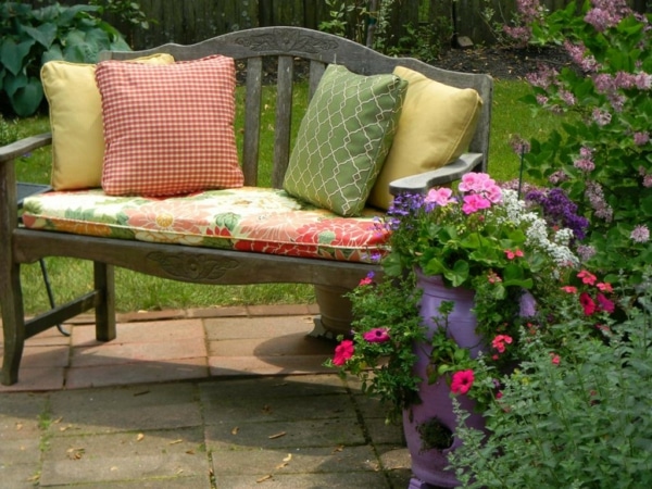 Banc de jardin tendance en bois avec quatre coussins dont deux jaunes, un vert et un autre rouge/blanc. Fleurs roses et rouges dans pot, pelouse en arrière-plan. crédit photo©Archzine