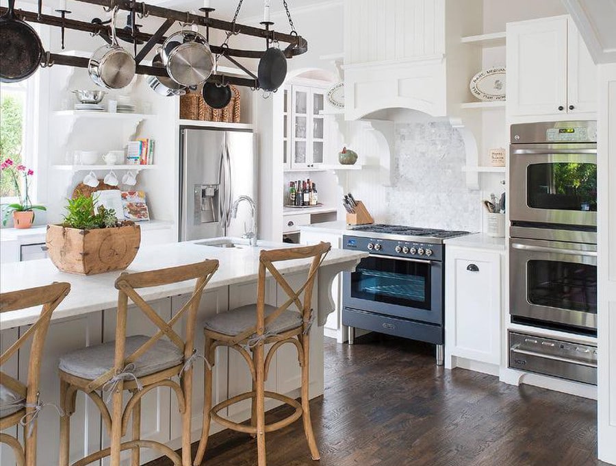 Cuisine avec un air de campagne donné grâce à des chaises en bois ainsi que casseroles et poêles suspendues au-dessus d'un plan de travail blanc. Parquet, four bleu-gris dans une salle plutôt blanche. Source : Ellen Grasso & Sons https://www.decorpad.com/designers/Ellen-Grasso-%26-Sons