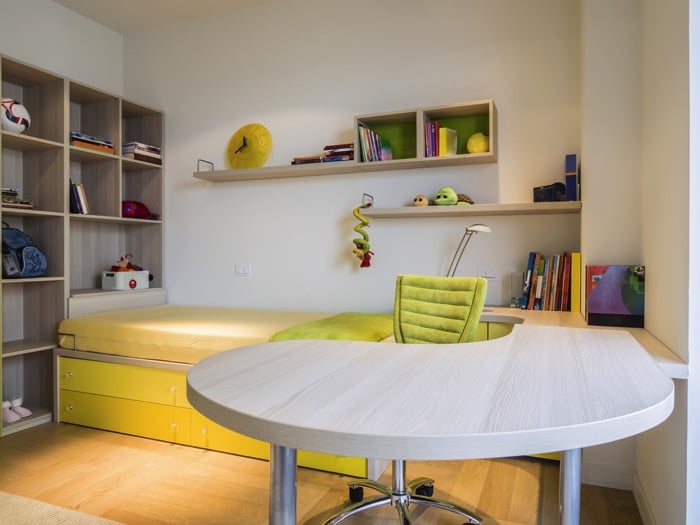 Chambre d'ado garçon originale décorée avec bureau blanc en L en bois. Parquet, chaise verte claire avec roulettes, lit une place, bibliothèque avec différentes cases, étagères avec peluches et horloge jaune dessus. deco-chambre-ado-garcon-copyright-basileus-0