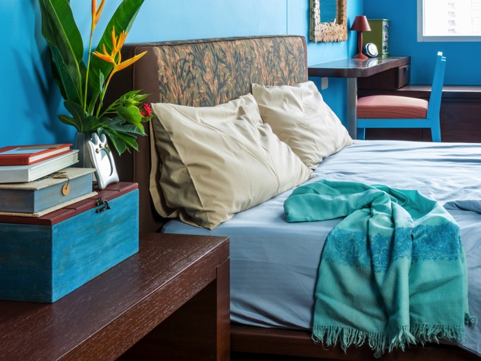 Chambre avec grand lit dans un style pop bleu clair avec deux bureaux en bois wengé. Miroir, petite lampes, fleurs dans un vase et rangements.
