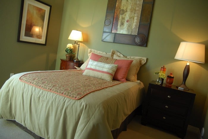 Chambre contemporaine composé de meubles en bois ainsi que de murs peints dans la couleur verte. Lit avec couette rose, blanche et beige avec deux tables de chevets différents et lampes blanches.