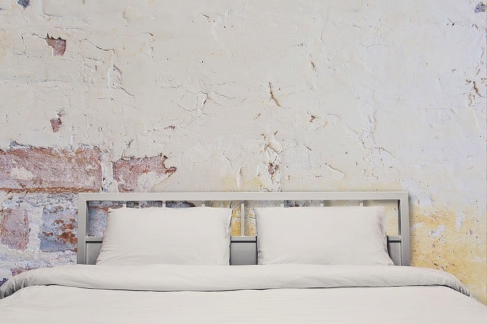 Chambre contemporaine brut avec grand lit blanc et en acier deux places avec coussins. Mur style industriel ancien en pierre.