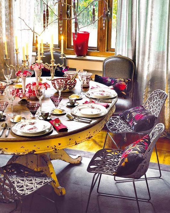 Salle à manger boho-chic avec table ovale en bois usé munie de différents fauteuils aux divers styles. Parquet, tapis gris et chandeliers en acier avec plusieurs bougies.