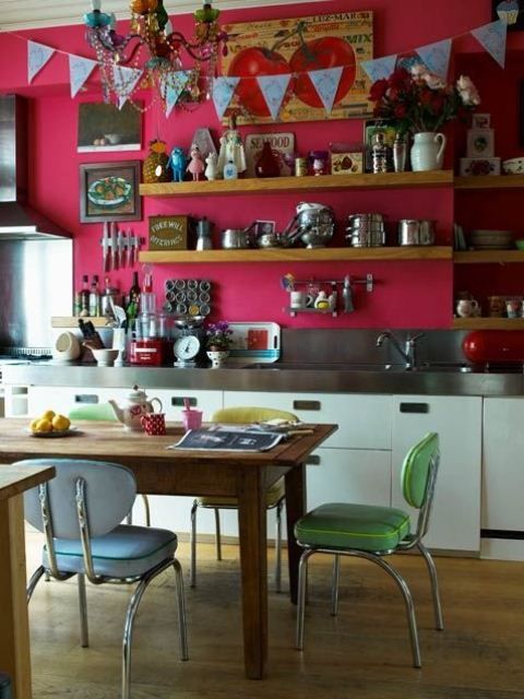 Salle à manger boho-chic avec table en bois sur parquet. Mur rose avec diverses étagères au-dessus d'un plan de travail en I où sont disposés une multitude de petits accessoires et tableau représentant des cerises.