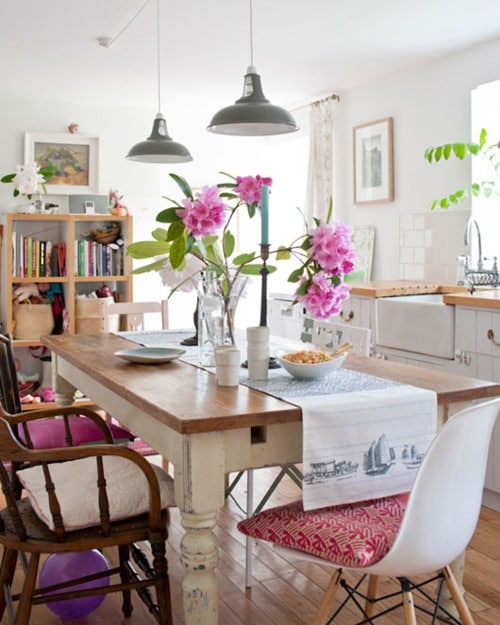 Salle à manger boho-chic moderne classique avec table en bois rectangulaire, pieds blancs, pot de fleurs roses. Petite bibliothèque en fond et deux lampes grises suspendues.