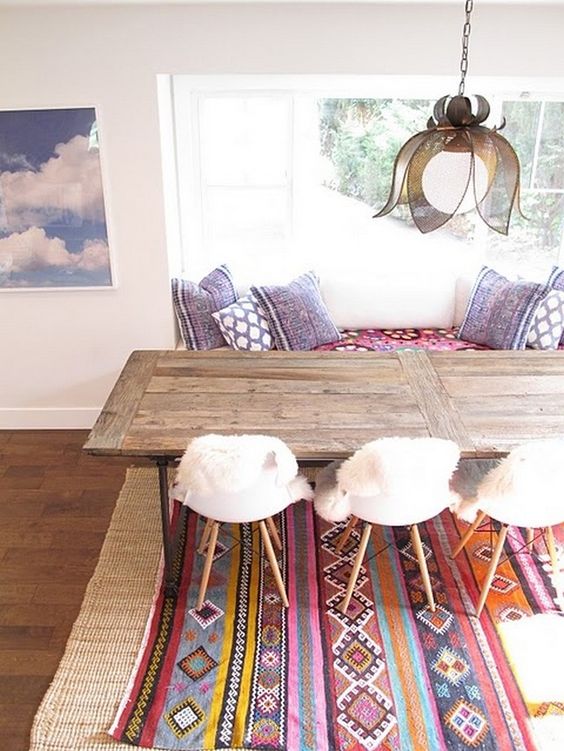 Salle à manger boho-chic avec table en bois rectangulaire dans une pièce toute blanche avec baie vitrée et tableau représentant le ciel sur la gauche. Tapis style africain.