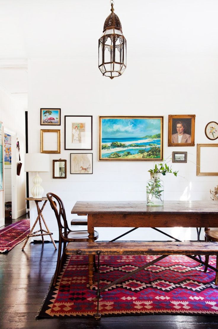 Salle à manger boho-chic blanche avec table en bois et tapis violet style africain. Multitude de tableaux dont des portraits et des œuvres naturalistes sur un mur totalement blanc.