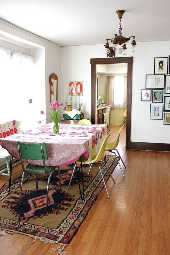 Salle à manger boho-chic dans une pièce blanche avec parquet stratifié. Table avec nappe rose sur tapis africain et multitude de petits tableaux différents sur la droite.