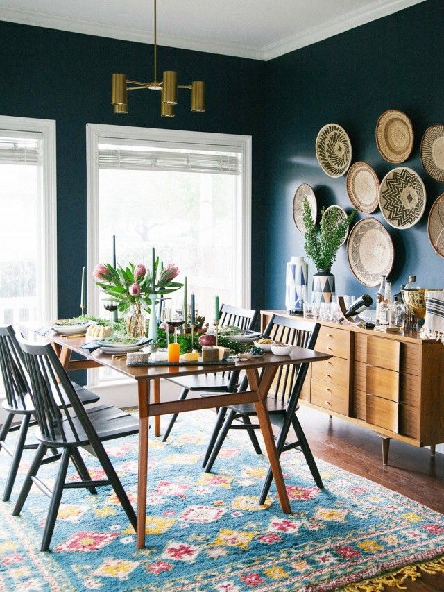 Salle à manger boho-chic avec table en bois rectangulaire sur tapis coloré original. Pièce avec mur bleu mer et plats tressés accrochés servant de décoration sur la droite.