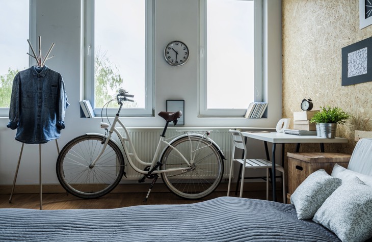 Bureau minimaliste moderne situé dans un coin d'une chambre et composée d'une table blanche, d'une chaise et d'un meuble de rangement en bois. Lit deux places, vélo, pendule et cadres sur mur.