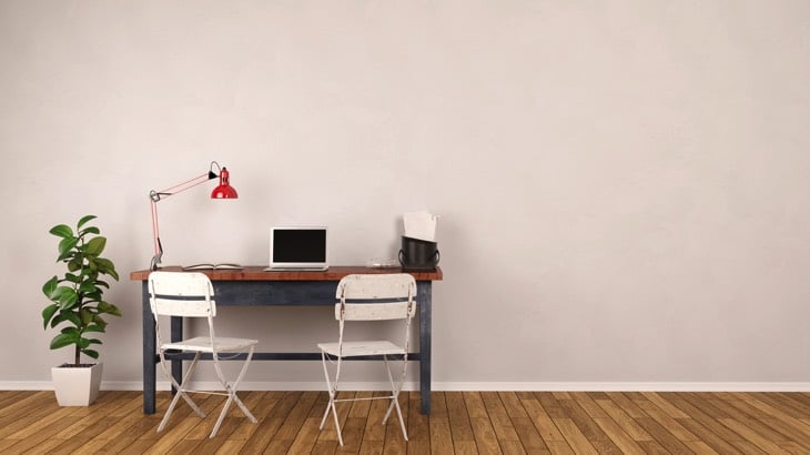 Bureau minimaliste placée au centre d'une pièce plutôt vide avec parquet et mur blanc.Deux chaises semblables, plantes dans pot, petit PC moderne et lampe rouge.