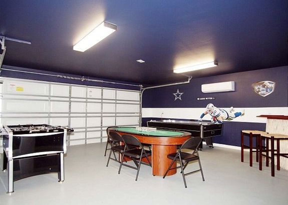 Garage original aménagé en salle de jeux bleu foncé et blanche avec grande porte de garage. Table de poker en bois, baby-foot, flipper et look rétro.