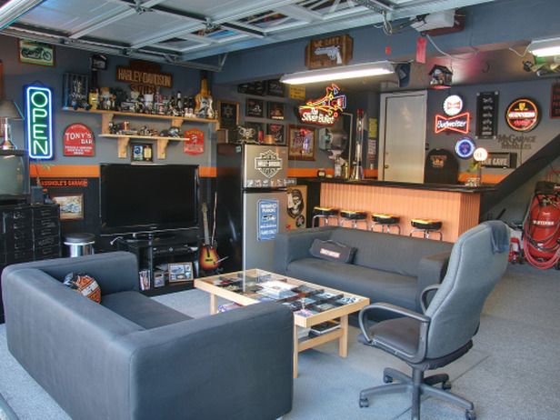 Garage aménagé en bar rétro original avec salon muni de fauteuils gris et d'une table en bois. TV, bouteilles, chaises de bar et frigo.