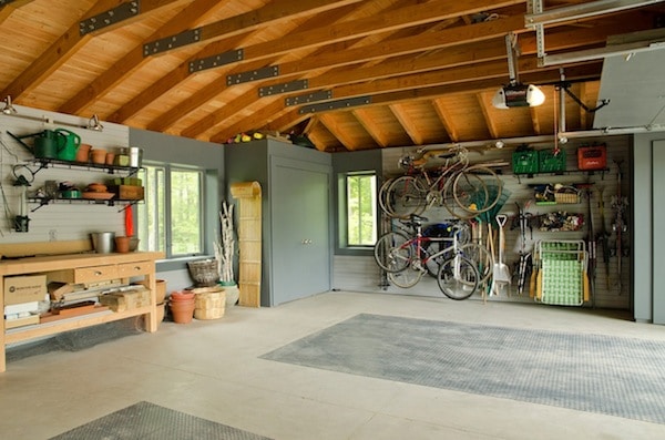 Garage aménagé avec plafond en bois et sol en carrelage. Établi en hêtre, étagères noires pour pots et arrosoirs ainsi que crochets afin de mettre des vélos.