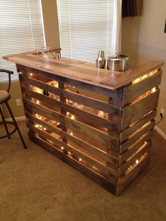 Bar en bois en U avec palettes espacées laissant apparaitre diverses ampoules. Verres et shakers dessus, moquette beige et deux stores blancs.