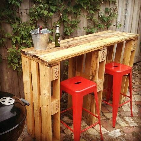 Bar en palettes rectangulaire extérieure assez petit avec deux tabourets plats carrés rouges. Petit seau avec différentes bières dedans, barbecue noir rond sur le côté.