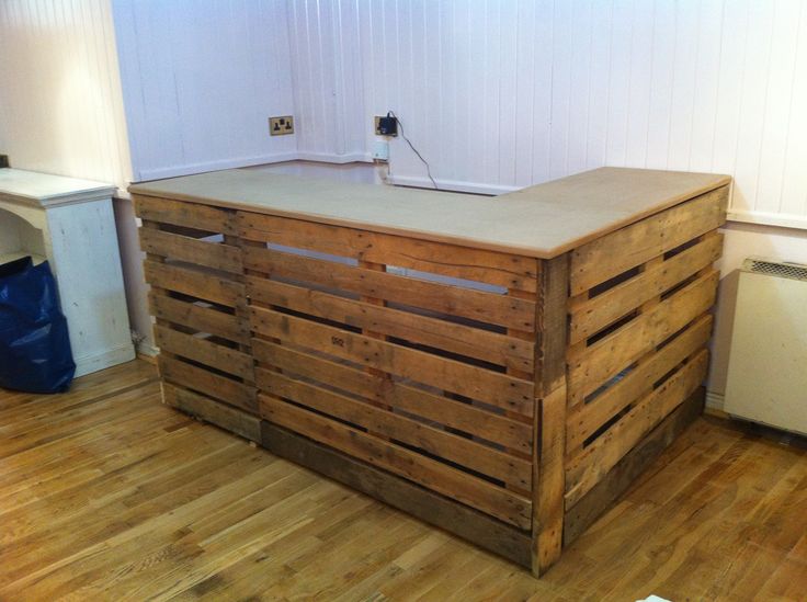 Bar en palettes bois vide avec dessus beige en L sur parquet stratifié. Pièce avec mur blanc avec cheminée vide blanche sur la gauche.
