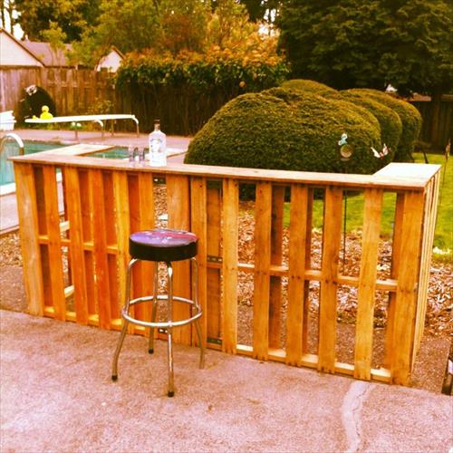 Bar en palette extérieur allongée en U avec un seul tabouret rond violet. Pelouse bien verte, grands buissons et piscine avec plongeoir et échelle à gauche.