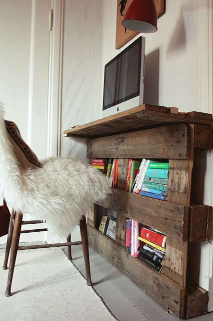 Bureau en palettes avec multitude de livres sur des rangements inférieurs. PC avec écran plat ultra moderne, tableau au-dessus et une chaise avec fourrure blanche.