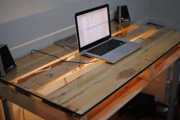 Bureau en palettes rectangulaire avec paroi de verre intégrée et un niveau de rangement inférieur. PC portable allumé et deux petites enceintes noires des deux côtés.
