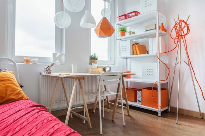 Déco ludique pour une chambre d’ado fille blanche avec parquet en chêne. Lit avec couverture rose et coussins oranges, table en bois et chaise blanche, lumières suspendues de différentes formes et rangements. chambre-ado-fille-photographee-eu-022