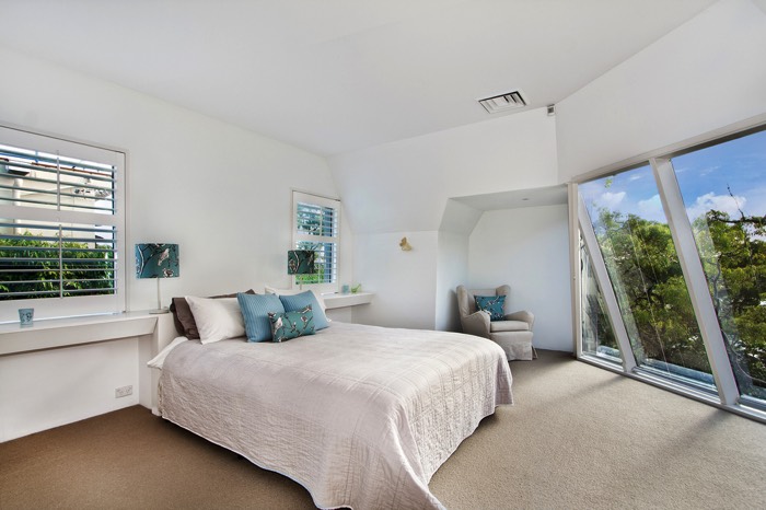 Chambre avec mur blanc, moquette beige, lit deux places et coussins en bleu clair. Petites fenêtres et grandes fenêtres en verre sur le côté.