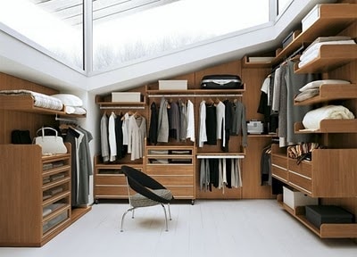 Dressing dans les combles en U en bois chêne servant pour vestes, chemises et costumes avec multitude d'étagères sur sol uni blanc. Plafond en verre.