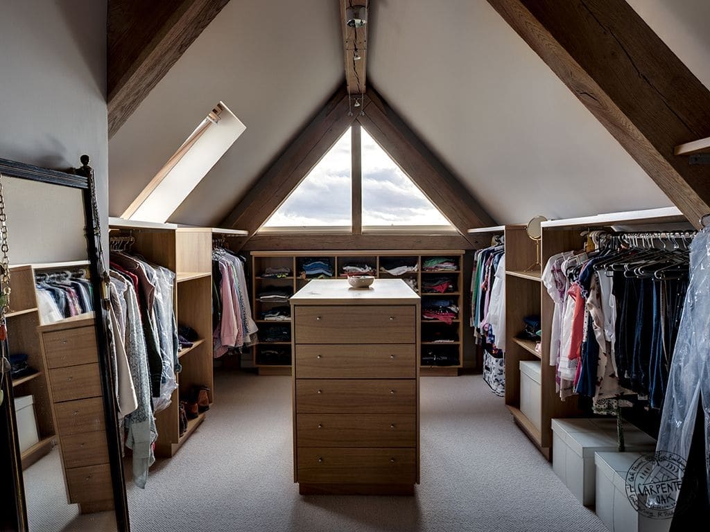 Dressing disposé en U dans les combles en bois chêne avec plafond beige et poutres. Meuble avec tiroirs sur moquette beige et miroir sur le côté gauche.