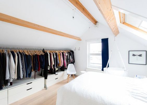 Dressing dans les combles avec chambre blanche style chalet et fenêtre avec rideau bleu foncé. Rangements sur parquet stratifié et grand lit blanc deux places.