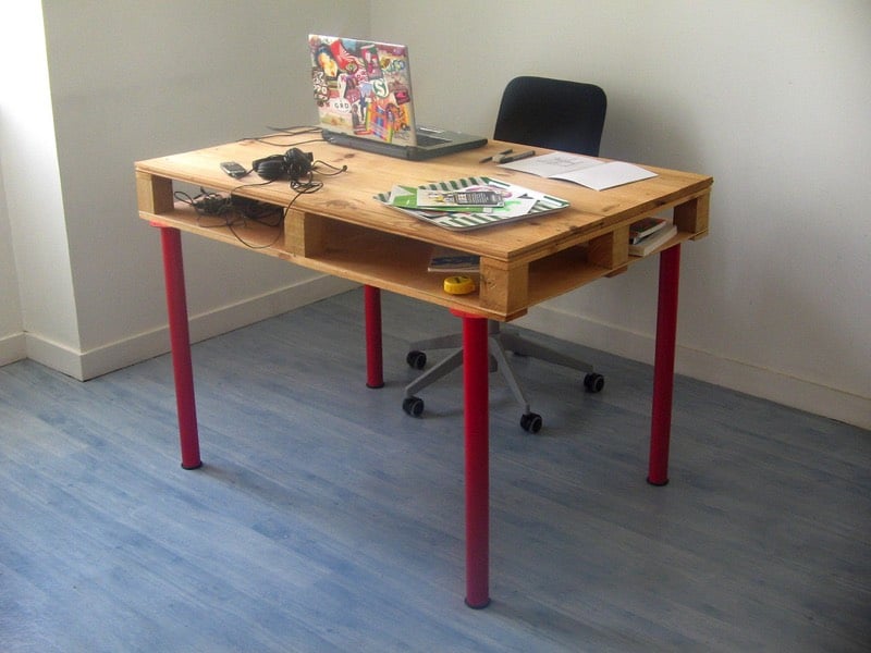 Petit bureau en palettes rectangulaire avec quatre pieds tout en rouge sur parquet et rangement en-dessous, siège classique noir muni de roulettes. PC avec dessin et graffitis multicolores au dos. fabriquer-bureau-en-palette-de-bois