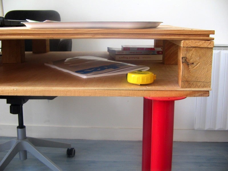 Bureau en palette doté d'un rangement inférieur avec magazine, mètre jaune et trois livres. Pieds rouges, chaise noire classique avec roulettes et parquet dans une pièce toute blanche. fabriquer-bureau-palette-bois-www-flickr-comphotos59716912n055931285682inphotostream