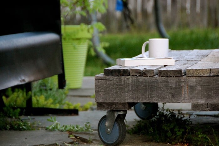 Table basse en bois munie de quatre roulettes bleues et noires, livre et tasse blanche dessus. Pot vert avec plantes, pelouse et canapé extérieur. fabriquer-table-basse-palette