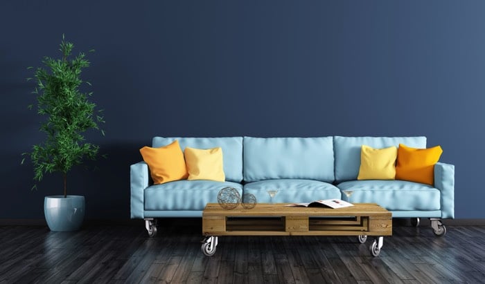 Fabriquer une table basse en bois chêne avec quatre petites roulettes sur parquet en bois foncé. Pot avec grande plante à gauche, canapé bleu clair avec coussins oranges et jaunes. fabriquer-table-basse-palette-vadym-andrushchenko-6
