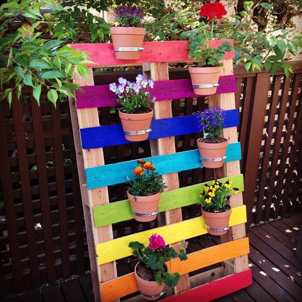 Jardinière en palette avec pot de fleurs contenant notamment des roses. Différentes peintures rose, violette, bleue foncé, bleue claire, vert, jaune, orange et rose.