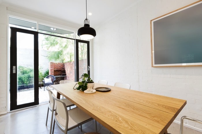 Table salle à manger en bois pin rectangulaire. Mur blanc avec portes noires coulissantes en verre donnant sur un extérieur avec vieux mur en briques rouges.