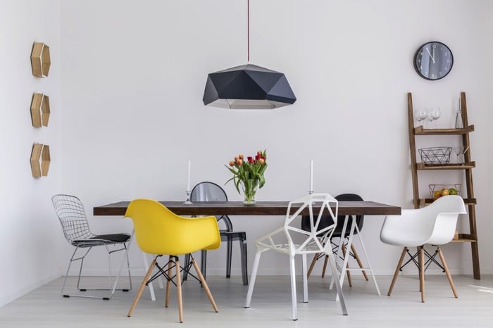 Table salle à manger en bois merbeau avec lampe bleue-gris suspendue. Plusieurs sièges au design original et de différentes couleurs dans une pièce au style minimaliste.