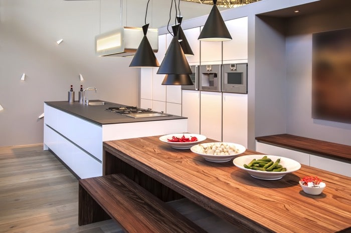 Table salle à manger en teck sur parquet stratifié. Cuisine blanche avec plan de travail en I gris, évier en inox et plaques de cuissons.