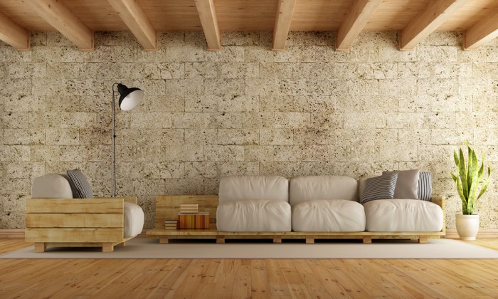 Fauteuil en palette avec coussins blancs sur tapis blanc rectangulaire et parquet hêtre. Mur en pierre, plantes sur la droite et lampe noire sur la gauche.