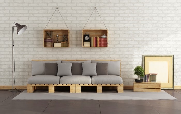 Fauteuil en palette avec coussins gris clair et gris foncé, miroir sur la droite et deux rangements suspendus en bois. Mur de pierres en blanc, tapis rectangulaire et carrelage.