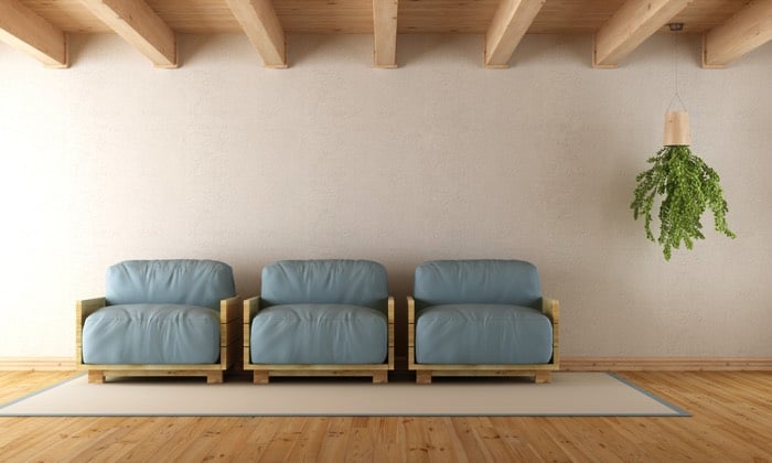 Fauteuil en palette avec coussins bleu-gris sur tapis beige avec ligne grise, mur uni. Parquet stratifié et plantes suspendue sur plafond en bois à droite. réalisation d'un fauteuil en palette de bois