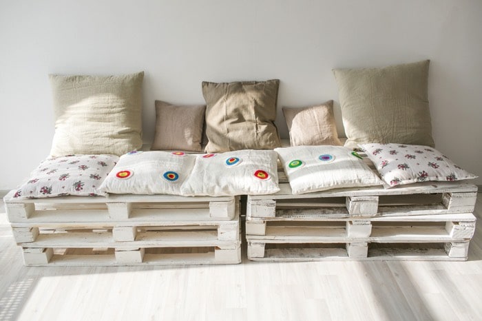 Fauteuils en palettes avec coussins de différentes couleurs gris, marron, beige et blancs avec divers motifs ronds multicolores. Mur blanc et parquet.