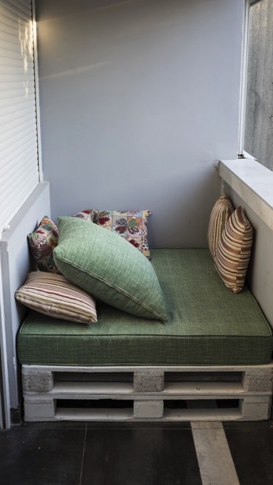 Fauteuil en palette avec des coussins verts, à rayures et avec des motifs. Mur blanc, carrelage noir et volet roulant blanc sur la gauche.