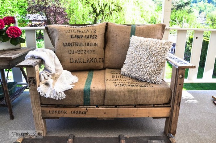 Fauteuil en palettes avec un coussin en laine et des coussins beiges avec inscriptions noires en anglais dessus. Terrasse avec clôture blanche et vue sur pelouse.