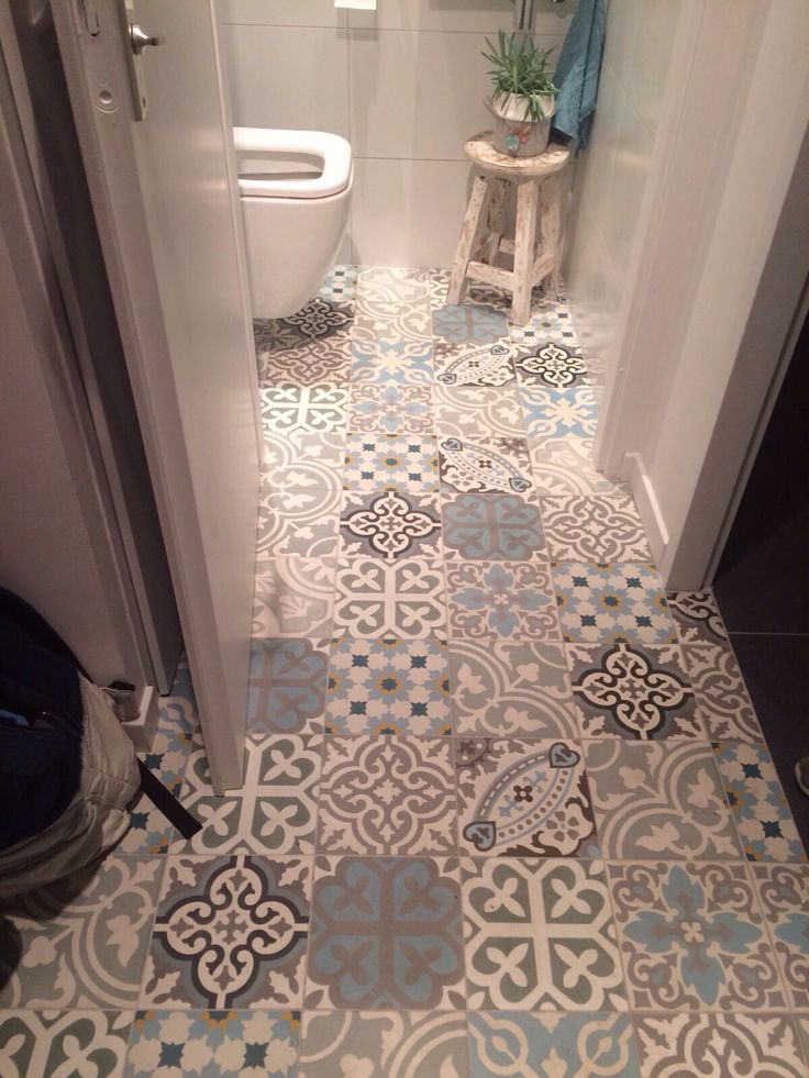 Carreaux de ciment à motifs arrondies de différentes couleurs dont le blanc, le gris ainsi que le bleu clair. Toilettes blanches avec tabouret en bois et plante dessus.