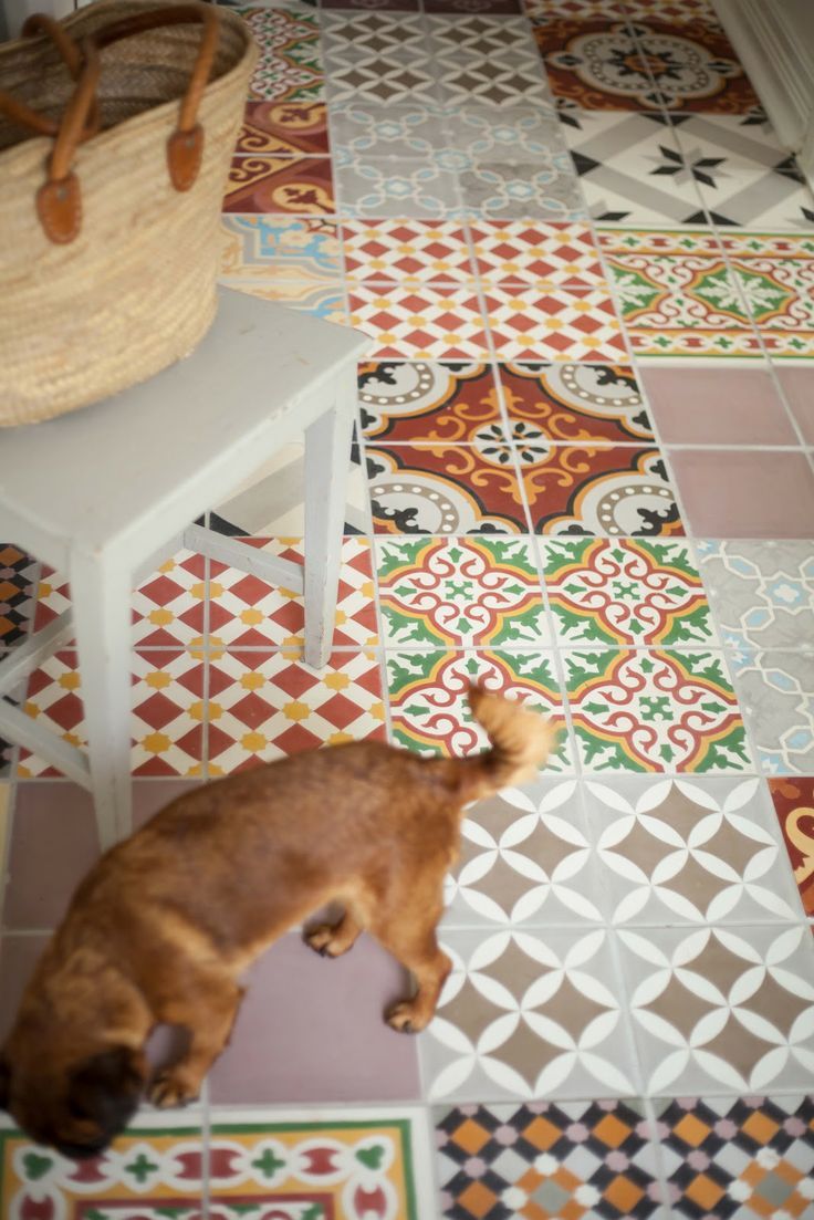 Carreaux en ciment de diverses formes avec des couleurs dont le rouge, le gris ainsi que le vert. Petite table rectangulaire blanche avec sac beige dessus.