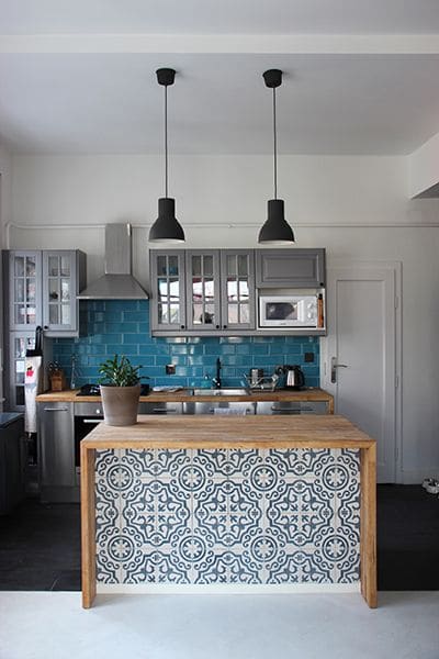 Carreaux de ciment blanc et bleu à motifs ronds et arrondis. Pièce avec deux lampes suspendues noires, cuisine avec plan de travail en bois hêtre avec carrelage mural bleu clair.