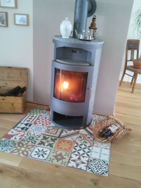 Carreaux en ciment de diverses formes et motifs avec des couleurs vertes, rouges et gris. Insert ovale gris avec foyer intérieur dans une pièce avec parquet.