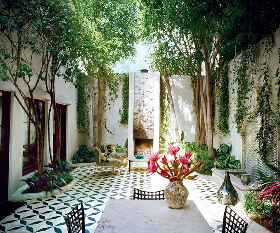 Carreaux en ciment noir et blanc dans une cour extérieur green power avec arbres, plantes grasses et lierre. Table à manger avec trois chaises noires et pot de fleurs roses dessus.