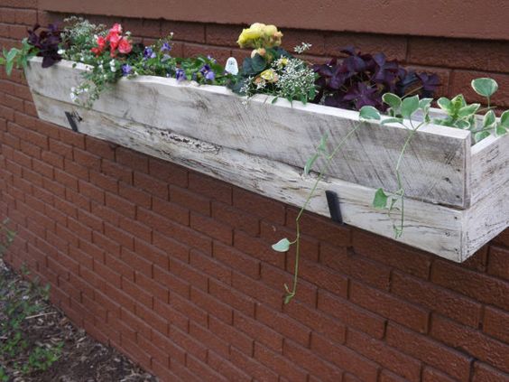 Jardinière en palette rectangulaire en bois blanc-gris avec multitude de fleur jaunes, roses et violettes dedans et de plantes. Mur en briques rouges.